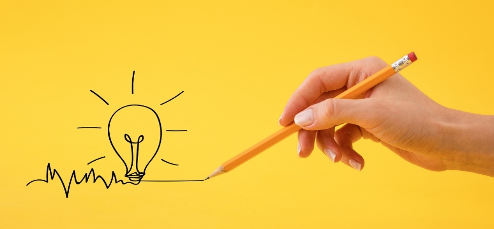 Hand holding a pencil drawing a glowing light bulb on a yellow background symbolizing creativity and ideas.