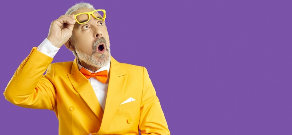 A man in a bright yellow suit and orange bow tie raises his glasses with a bewildered expression against a plain purple background.